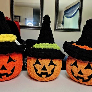 May include: Three crocheted orange pumpkins with black jack-o'-lantern faces and black witch hats. The hats feature textured bands in yellow, lime green, and orange. Each pumpkin has stitched black eyes with eyelashes and a wide, toothy grin.