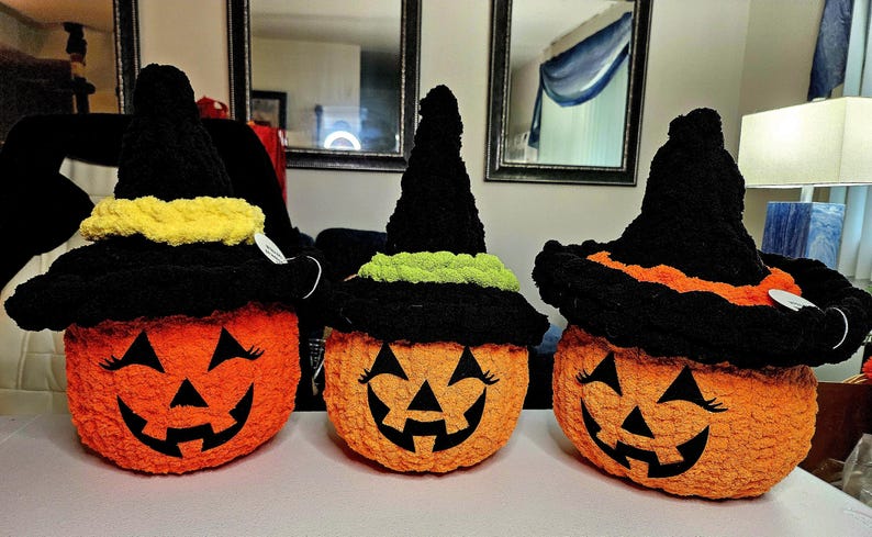 May include: Three crocheted orange pumpkins with black jack-o'-lantern faces and black witch hats. The hats feature textured bands in yellow, lime green, and orange. Each pumpkin has stitched black eyes with eyelashes and a wide, toothy grin.