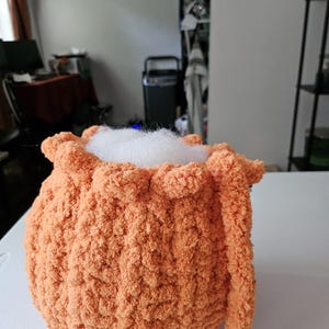 May include: A round, orange, crocheted plush object with a textured, fuzzy yarn surface. The top opening is gathered with a ruffled edge and is filled with white, fluffy stuffing material that protrudes slightly from the center of the item.