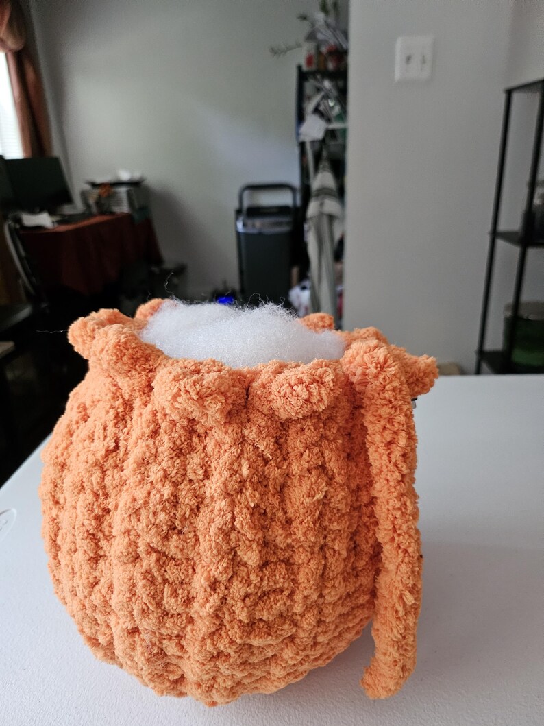 May include: A round, orange, crocheted plush object with a textured, fuzzy yarn surface. The top opening is gathered with a ruffled edge and is filled with white, fluffy stuffing material that protrudes slightly from the center of the item.