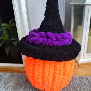 May include: A crocheted pumpkin figure featuring a bright orange base and a black pointed witch hat. The hat is accented with a thick, textured purple band around the brim. The entire object is crafted from soft, chunky yarn with a visible knit texture.