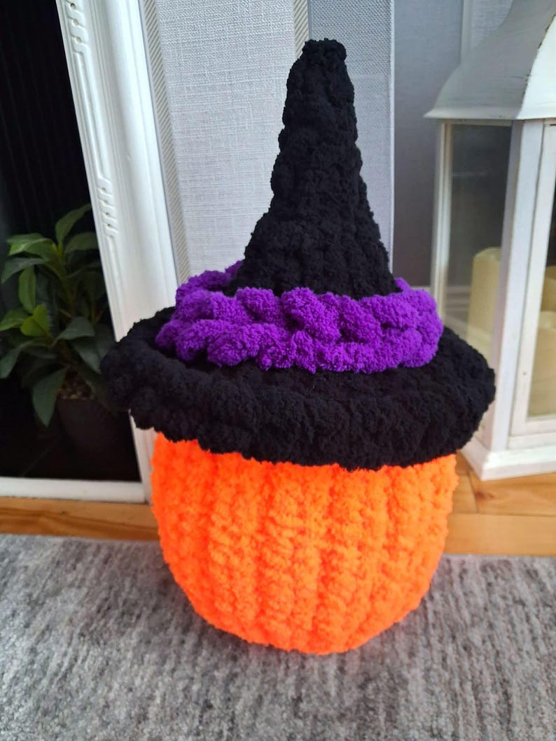 May include: A crocheted pumpkin figure featuring a bright orange base and a black pointed witch hat. The hat is accented with a thick, textured purple band around the brim. The entire object is crafted from soft, chunky yarn with a visible knit texture.