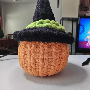 May include: A plush, crocheted pumpkin figure featuring a textured orange body and a black pointed witch hat. The hat is accented with a band of bright green yarn around the base. The entire object is crafted from soft, fuzzy yarn material.