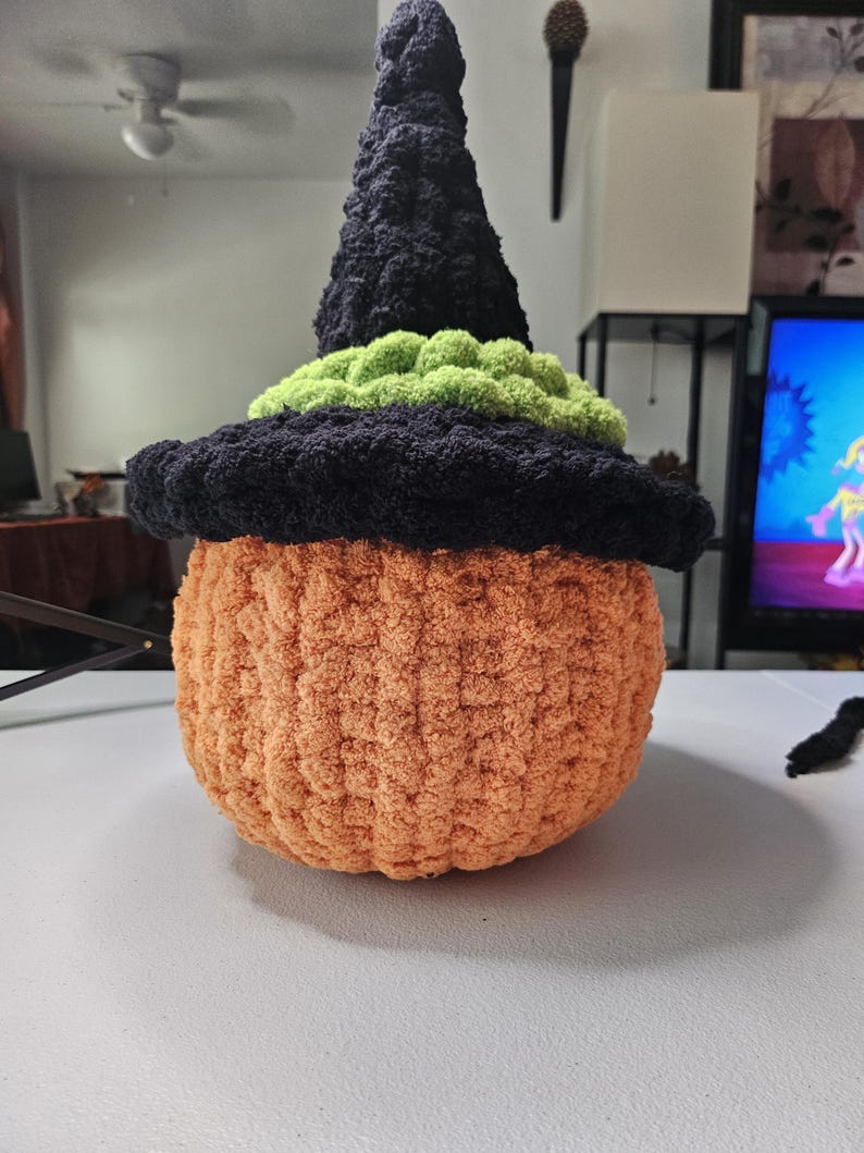 May include: A plush, crocheted pumpkin figure featuring a textured orange body and a black pointed witch hat. The hat is accented with a band of bright green yarn around the base. The entire object is crafted from soft, fuzzy yarn material.