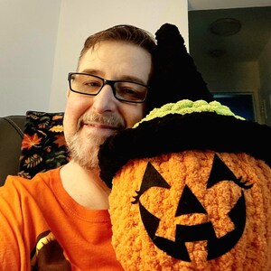 May include: A plush, round, orange pumpkin toy with a textured, fuzzy surface. It features a black felt jack-o'-lantern face with triangular eyes and a jagged mouth. The pumpkin is topped with a black and lime green crocheted witch hat.