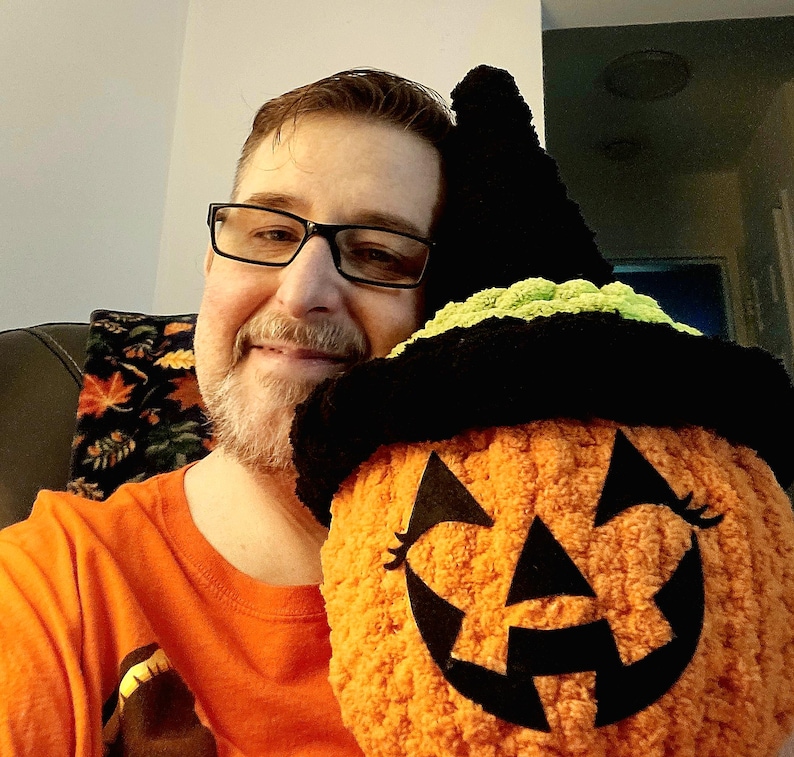 May include: A plush, round, orange pumpkin toy with a textured, fuzzy surface. It features a black felt jack-o'-lantern face with triangular eyes and a jagged mouth. The pumpkin is topped with a black and lime green crocheted witch hat.