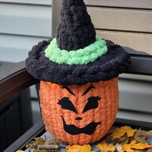 May include: A crocheted orange pumpkin figure wearing a black witch hat with a bright green band. The pumpkin has a black felt face with arched eyebrows, almond-shaped eyes, and a wide, curved mouth. The texture is soft, fuzzy, and yarn-based.