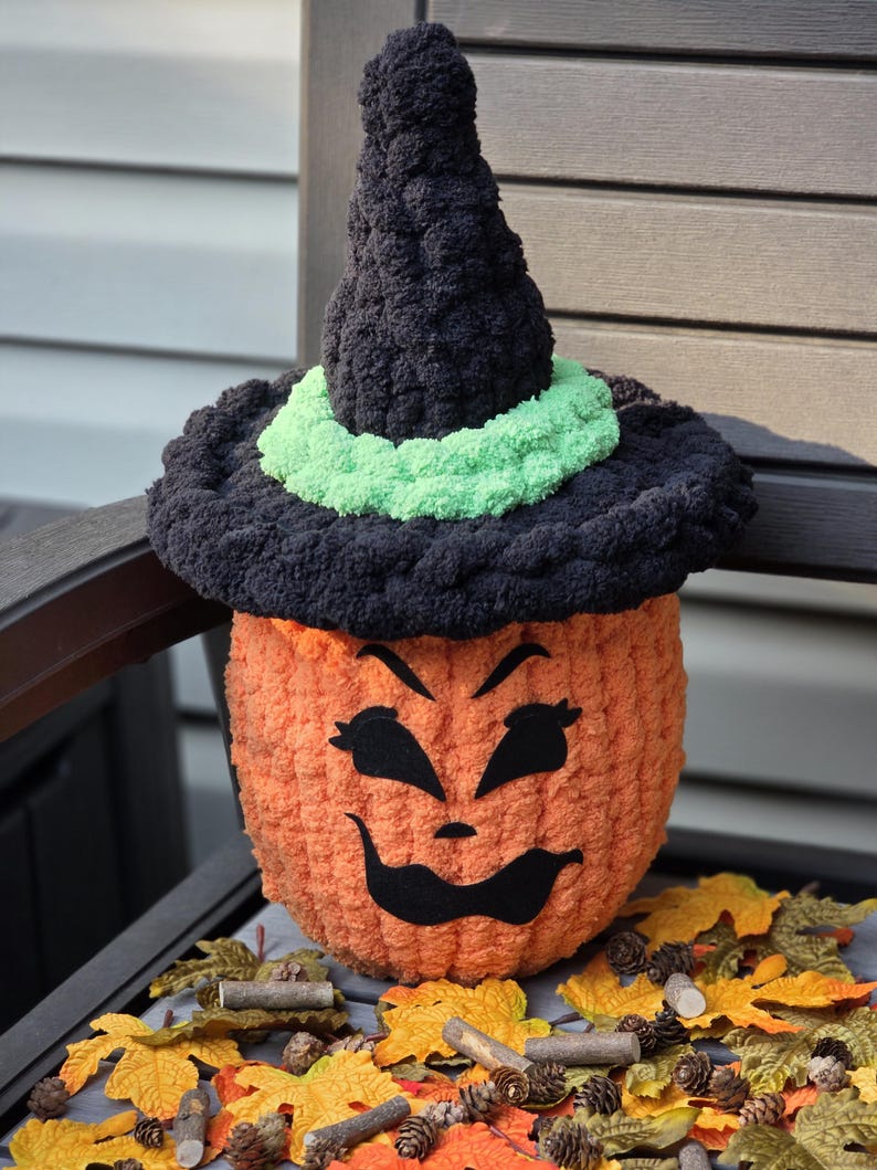 May include: A crocheted orange pumpkin figure wearing a black witch hat with a bright green band. The pumpkin has a black felt face with arched eyebrows, almond-shaped eyes, and a wide, curved mouth. The texture is soft, fuzzy, and yarn-based.