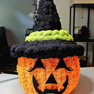 May include: A round, orange, textured yarn pumpkin wearing a tall, black, crocheted witch hat with a bright lime green band. The pumpkin features a black felt jack-o'-lantern face with triangular eyes, long eyelashes, and a jagged mouth.