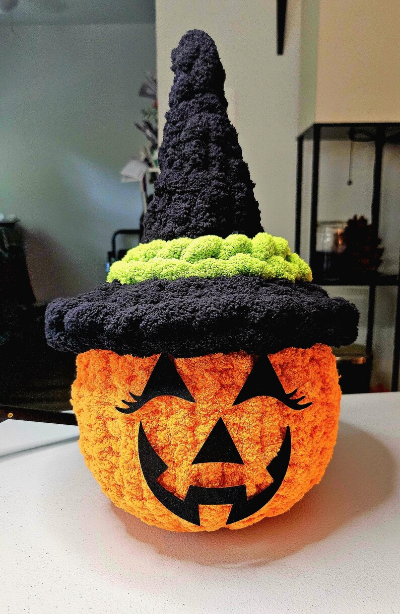 May include: A round, orange, textured yarn pumpkin wearing a tall, black, crocheted witch hat with a bright lime green band. The pumpkin features a black felt jack-o'-lantern face with triangular eyes, long eyelashes, and a jagged mouth.