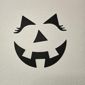 May include: A set of black felt cutouts forming a jack-o'-lantern face. The pieces include two triangular eyes with long eyelashes, a small triangular nose, and a wide, curved mouth with two square teeth pointing downward from the top edge.