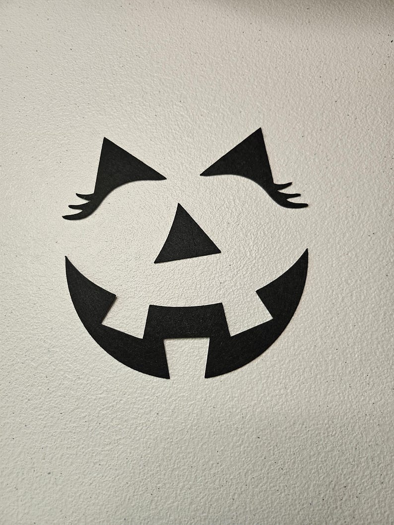 May include: A set of black felt cutouts forming a jack-o'-lantern face. The pieces include two triangular eyes with long eyelashes, a small triangular nose, and a wide, curved mouth with two square teeth pointing downward from the top edge.