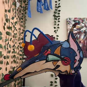 May include: A handcrafted mask featuring a stylized creature head with a long, pointed snout, blue and cream coloring, and dark outlines. It includes two blue pipe cleaner antennae topped with yellow pom-poms and a maroon-colored hair element.