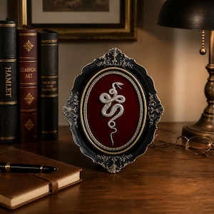 May include: An oval-shaped decorative frame with an ornate black and gold border, featuring a silver-toned coiled snake relief centered on a deep red velvet background. The frame has intricate scrollwork details along its outer edges.