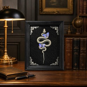 May include: A rectangular black frame containing a white snake skeleton mounted on a black textured background. Two small blue butterflies are positioned on the snake's body. The frame features ornate silver-toned metal corner accents.