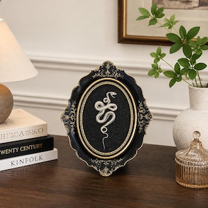 May include: An oval-shaped decorative frame with a black and gold finish featuring an intricate, ornate border. Inside the frame, a silver-toned snake figure is displayed against a textured black background, creating a striking contrast.