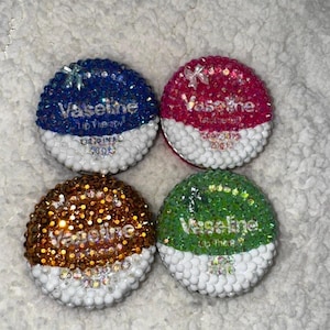 May include: Four circular Vaseline Lip Therapy containers, each decorated with small, colourful rhinestones. The top halves feature blue, pink, gold, and green crystals, while the bottom halves are covered in white beads, displaying the 20g weight.