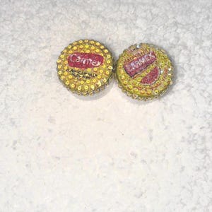 May include: Two circular Carmex lip balm containers featuring yellow lids adorned with small, reflective, gold-toned circular embellishments. The lids display the red and white Carmex logo and text, with one lid showing the words Classic Lip Balm Medicated.