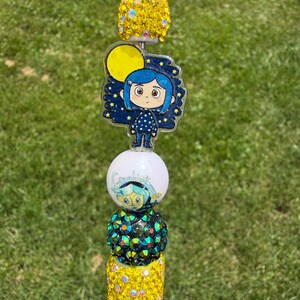 Puede incluir: Un adorno para bol&iacute;grafo con una esfera de diamantes de imitaci&oacute;n amarillos, un dije acr&iacute;lico plano de un personaje de pelo azul sobre un cielo estrellado con una luna amarilla, una cuenta blanca con una cara impresa, una esfera verde oscuro.