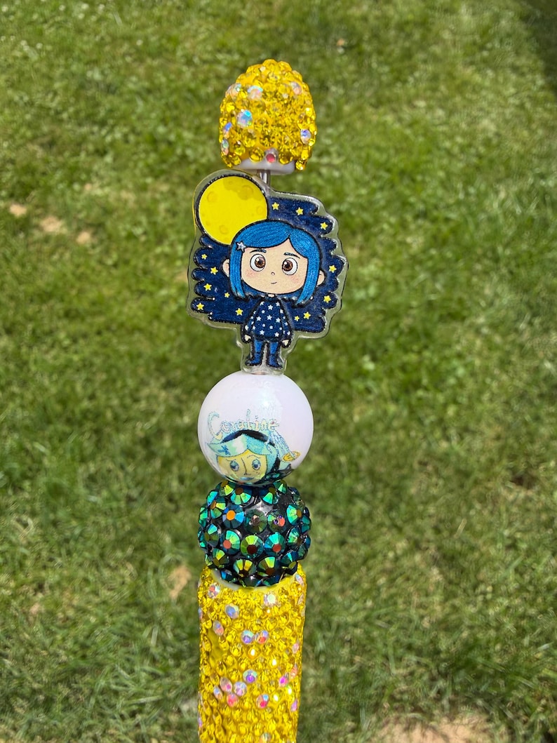 Puede incluir: Un adorno para bol&iacute;grafo con una esfera de diamantes de imitaci&oacute;n amarillos, un dije acr&iacute;lico plano de un personaje de pelo azul sobre un cielo estrellado con una luna amarilla, una cuenta blanca con una cara impresa, una esfera verde oscuro.
