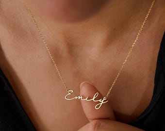 May include: A gold-toned metal necklace featuring a delicate chain and a script-style nameplate pendant that reads Emily. The pendant is attached to the chain on both sides, creating a centered display of the name in a cursive font style.