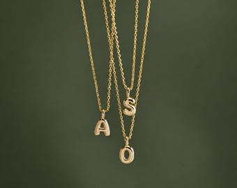 Puede incluir: Tres collares dorados, cada uno con un colgante de letra pulida. Un collar muestra la letra A, otro la letra S y el tercero la letra O. Las cadenas son finas y metálicas, colgando verticalmente sobre un fondo oscuro.