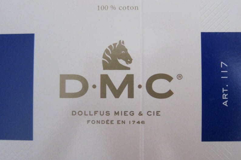 May include: A white label with a gold logo for DMC, a company that makes embroidery floss. The logo features a stylized horse's head and the text "D.M.C.  DOLLFUS MIEG & CIE FOND&Eacute;E EN 1746". The label also has the text "100% coton" and "ART. 117" in blue.