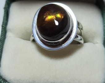 May include: A silver ring with a large, oval, dark brown gemstone. The gemstone has internal golden and yellow flashes. The ring is set in a silver bezel and sits in a green velvet ring box.
