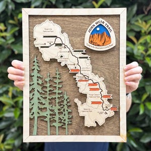 May include: A wooden wall art piece featuring a map of the John Muir Trail. The map includes a light wood cutout of the trail path, green pine tree silhouettes, and a circular badge at the top with the text John Muir Trail and Going to the Mountains is Going Home.