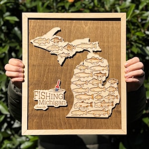 May include: A rectangular wooden wall art piece featuring a laser-cut map of Michigan with various fish species engraved on the surface. The text Fishing Michigan is displayed with a red and blue fishing bobber icon on a light wood cutout.