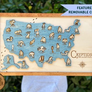 May include: A wooden map of the United States with a blue finish, featuring 33 removable wooden cryptid figures placed across various states. The map includes the text "Cryptids of the united states" and a compass rose, all enclosed in a wooden frame.