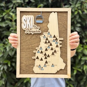May include: A rectangular wooden wall art piece featuring a map of New Hampshire with the text "SKI New Hampshire" and a light blue ski lift icon. The map includes numerous small dark brown and light blue mountain icons labeled with ski resort names.