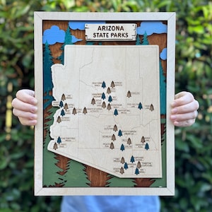 Peut inclure: Une carte en bois encadrée des parcs d'État de l'Arizona avec un cadre en bois clair, un fond en bois foncé avec des nuages bleus superposés et des pins verts, et une carte centrale avec des icônes d'arbres marquant les parcs.
