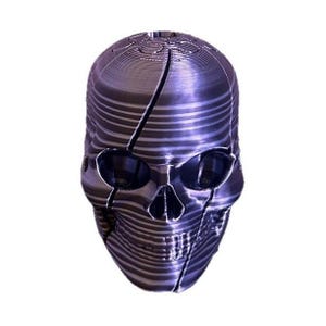 May include: A 3D printed skull object with a metallic purple finish and visible horizontal layer lines. The skull features deep, hollow eye sockets, a nasal cavity, and a defined jawline with a prominent crack running vertically across the face.