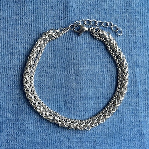 Può includere: Un bracciale a catena multistrato color argento composto da una spessa catena intrecciata a spiga e una catena più sottile a palline. Il bracciale presenta una chiusura a moschettone e una catenina di estensione regolabile con maglie tonde.
