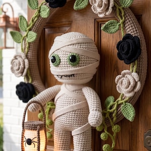 Könnte beinhalten: Eine gehäkelte Mumienpuppe, die eine kleine beleuchtete Laterne mit einer schwarzen Spinnengrafik hält, befestigt an einem runden Kranz, der mit gehäkelten schwarzen und beigen Rosen, grünen Blättern und Ranken verziert ist, hängend an einer Holztür.