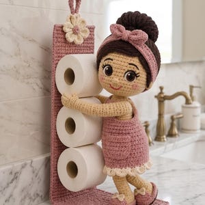 Könnte beinhalten: Ein gehäkelter Toilettenpapierhalter mit einer Puppe mit dunkelbraunem Dutt, die ein rosa Kleid und ein Stirnband trägt. Die Puppe ist an einer vertikalen rosa Basis mit einer cremefarbenen Blume befestigt und hält drei weiße Papierrollen.
