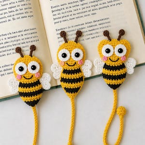 Könnte beinhalten: Drei handgefertigte Häkel-Lesezeichen in Bienenform mit gelb-schwarz gestreiften Körpern, weißen Flügeln, großen weißen Augen mit schwarzen Pupillen, rosa Wangen und braunen Fühlern. Jede Biene hat einen langen gelben Schwanz, der in einer kleinen gelben Kugel endet.