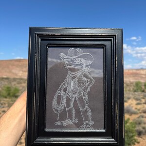 May include: A black rectangular picture frame containing a white line art illustration of a frog dressed as a cowboy. The frog wears a wide-brimmed hat, a neckerchief, a vest, chaps, and spurs, while holding a coiled lasso in one hand.