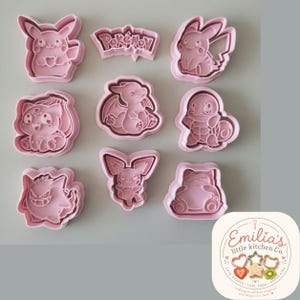 May include: A set of eight light pink plastic cookie cutters featuring various character shapes and a logo plaque with the text Pokémon. The collection includes designs of popular creatures, each with detailed outlines for pressing into dough.