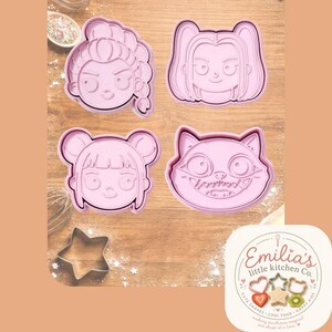 May include: A set of four light pink plastic cookie cutters featuring three distinct human face designs with different hairstyles and one cat face design. A separate metal star-shaped cookie cutter is included in the set. The brand logo Emilia's little kitchen Co. is visible in the corner.