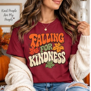 May include: A maroon short-sleeved t-shirt featuring a graphic design with the text "FALLING FOR KINDNESS" in a retro font. The design includes orange, yellow, and green maple leaf illustrations interspersed among the bold, wavy lettering.