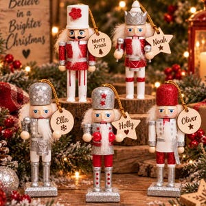 May include: Five wooden nutcracker ornaments featuring red, white, and silver glitter finishes. Each figure holds a small drum or staff and includes a wooden tag with names like Noah, Ella, Holly, and Oliver, or the text Merry Xmas with a tree icon.
