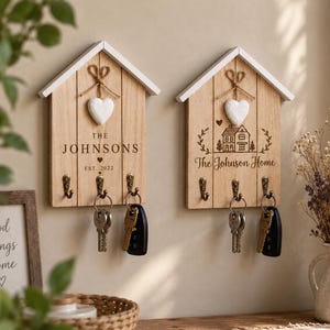 May include: Two house-shaped wooden key holders with white roofs and three metal hooks each. The left holder features the text THE JOHNSONS EST. 2022, while the right displays a house illustration and The Johnson Home text. Both have a white heart.