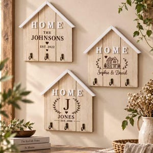 May include: Three house-shaped wooden wall organisers with white gabled roofs. Each features the word HOME in white letters, three black metal hooks, and personalised text including The Johnsons Est. 2022, a house illustration with Sophie & David, and J Jones Est. 2008.