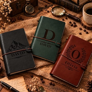 May include: Three rectangular journals in black, dark green, and brown with elastic closures. The black journal features a mountain graphic and the name DAVID. The green journal shows a large letter D and DAVID. The brown journal displays a letter O with a wreath and the name Olivia.