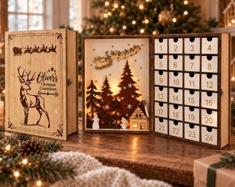 Könnte beinhalten: Ein hölzerner Adventskalender in Buchform mit einem Hirsch und dem Text "Oliver's Christmas Countdown" auf dem Einband. Das Innere zeigt eine hinterleuchtete Waldszene mit Haus, Schneemann und Weihnachtsmannschlitten sowie vierundzwanzig nummerierte Schubladen.