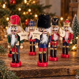 May include: A collection of wooden nutcracker ornaments featuring various uniforms in red, green, blue, and black. Each figure has a white beard and a hanging tag with text including Myla, Missing you, and Lily, suspended by gold strings for display.
