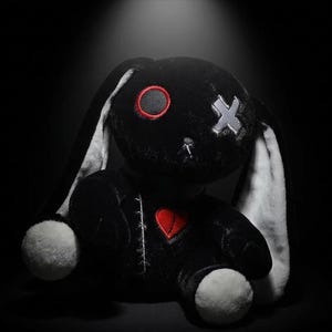 May include: A plush rabbit toy featuring black fabric with long white inner ears and white paws. The face has one circular red eye and one grey X-shaped eye. A red heart is stitched onto the chest, and white stitching details run along the body.