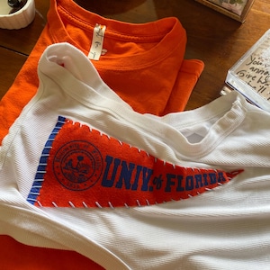 May include: A white ribbed tank top featuring an orange felt pennant patch with blue text that reads Univ. of Florida and a circular seal. Beneath the tank top is a solid orange t-shirt with a tag that reads Just for you.